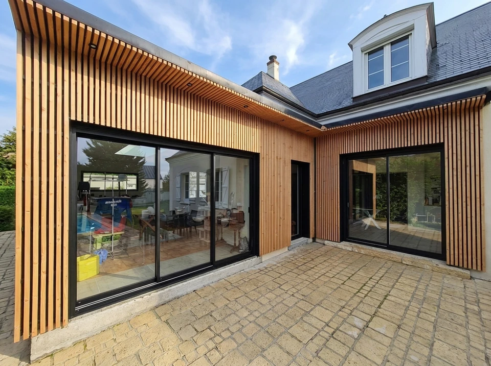 Intérieur d'un showroom de menuiserie moderne et accueillant avec des fenêtres PVC et aluminium, des portes d'entrée et des baies vitrées exposées. Un menuisier professionnel discute avec un couple de clients dans le showroom, pointant des détails de produits de menuiserie. La lumière naturelle illumine les différents modèles et finitions de fenêtres et portes exposés. L'atmosphère est professionnelle et rassurante, mettant en avant l'expertise et l'accompagnement personnalisé du menuisier auprès de ses clients.